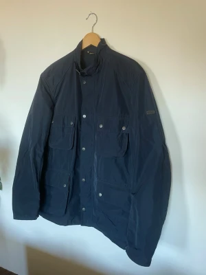 Barbour International Weir Casual Field Jacket – Marinblå XL - Säljer en stilren och klassisk field jacket från Barbour International i modellen Weir Casual. Jackan är i en snygg marinblå färg och är den perfekta lättviktsjackan för vår, sommar och tidig höst. Inspirerad av den klassiska motorcykelstilen med fyra utanpåliggande fickor, hög krage med spänne, kviltade förstärkningar vid armbågarna och Barbour-logotyp på ärmen. En tidlös modell som passar till det mesta!  Bild 1-3 har mest korrekt färgåtergivning. Bild 4-7  visar skönhetsfel, som beskrivs neda