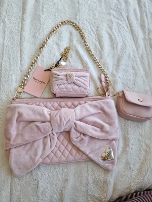 Rosa quiltad handväska Juicy Couture - Supersöt ljusrosa handväska från Juicy Couture i quiltad sammet med stor rosett framtill. Väskan har guldfärgad kedjerem, matchande liten plånbok och miniväska, samt hjärtformad Juicy Couture-detalj. Perfekt för dig som älskar gulliga accessoarer!