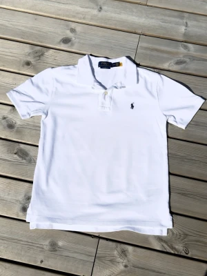 Vit pikétröja från Polo Ralph Lauren - Klassisk vit pikétröja från Polo Ralph Lauren med korta ärmar och krage. Tröjan har knappar framtill och den ikoniska broderade loggan på bröstet. Perfekt till jeans eller shorts för en clean och stilren look.