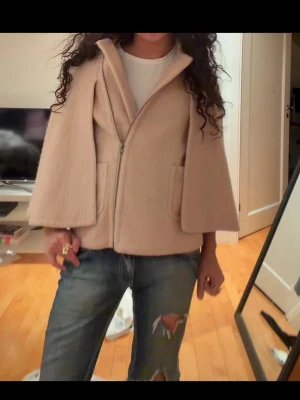 Beige capejacka med dragkedja - Snygg beige capejacka med dragkedja framtill. Jackan har två stora fickor och en uppvikt krage. Tillverkad i ett mjukt, ull-liknande material som ger en lyxig känsla. Perfekt för dig som vill ha en trendig och unik look.