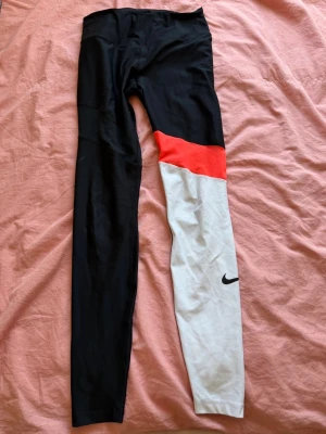Nike leggings med färgblock - Snygga leggings från Nike med unik färgblockdesign: ena benet är helsvart, det andra är svart upptill, rött på mitten och vitt nedtill med en svart Nike-logga. Perfekta för träning eller chill. Stretchigt material för skön passform.