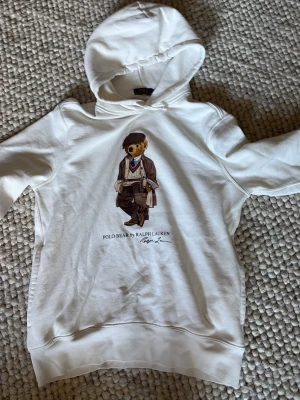 Vit hoodie Polo Bear Ralph Lauren - Snygg vit hoodie från Ralph Lauren med den ikoniska Polo Bear-trycket på bröstet. Klassisk huva och ribbade muddar. Perfekt för dig som gillar streetwear och vill ha en stilren look med en twist. Materialet känns mjukt och skönt mot huden.