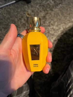 Erba Gold parfym - Snygg parfymflaska från Xerjoff i en klargul färg med gulddetaljer och elegant lock. Flaskan har en modern, rundad form och en metallisk etikett framtill där det står 'Erba Gold'. Perfekt för dig som gillar lyxiga och unika dofter. Ej använd.