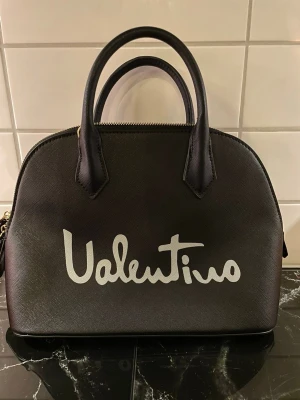Svart handväska från Valentino - Snygg svart handväska från Valentino med vit logga framtill. Väskan har rundad form, dubbla handtag och avtagbar axelrem i läder. Guldiga metalldetaljer och dragkedja upptill. Perfekt för dig som gillar stilrena accessoarer med statement.
