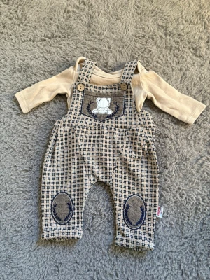 Beige babyset med hängselbyxor - Sött babyset med beige långärmad tröja och rutiga hängselbyxor i beige och mörkblått. Byxorna har broderad björn på bröstet och ovala knäpatchar. Mjukt material, perfekt för små barn. Använd endast en gång 