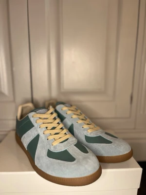  ljusblå och grön/mörkblå sneakers Maison Margiela - Snygga sneakers från Maison Margiela i grönt och ljusblått med beige snören och gummisula. Ovandelen är i mocka och läder, klassisk låg modell med retrovibbar. Perfekta för dig som gillar stilrena detaljer och unik design. Can sänka priset