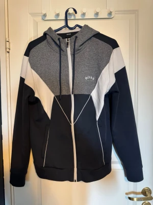 Hugo boss Tracksuit - Snyggt loungewear set från BOSS med zip-hoodie och matchande byxor. Setet är i svart, grått och vitt med coola geometriska paneler och BOSS-logga på både tröja och byxa. Hoodien har dragkedja och fickor, byxorna har dragsko i midjan. Perfekt för chill dagar hemma.