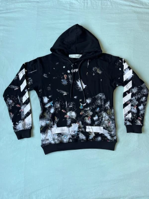 Off-White svart hoodie med tryck - Svart hoodie från Off-White med färgstänk och vit grafisk design på ärmarna. Klassiska snören i huvan och ribbade muddar. Unik streetwear-look med Off-White-logga och detaljer i vitt och flera färger. Perfekt för dig som gillar statement-plagg.