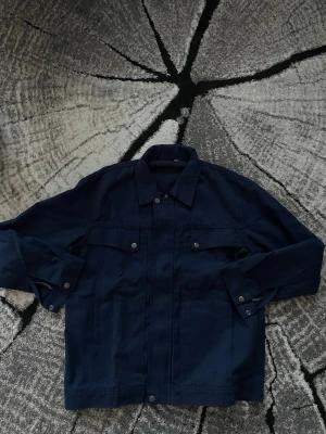 Mörkblå jeansjacka med fickor - Snygg mörkblå jeansjacka med klassisk krage och två bröstfickor med knappar. Jackan har raka ärmar och silverfärgade knappar framtill. Perfekt för dig som gillar en stilren och tidlös look.