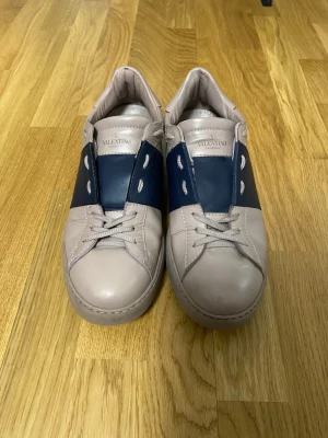 Valentino skor - Snygga sneakers från Valentino i ljusbeige läder med en bred mörkblå läderdetalj över mitten. Skorna har klassisk snörning och diskreta nitar längs sulan för en cool touch. Perfekta för dig som gillar stilrena men unika sneakers.