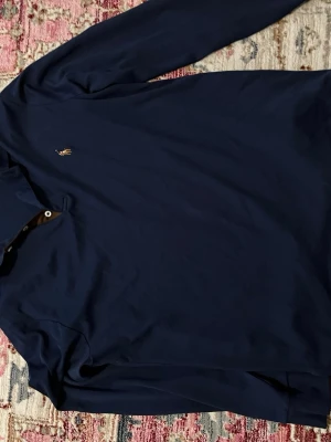 Mörkblå långärmad skjorta Polo Ralph Lauren - Mörkblå långärmad skjorta från Polo Ralph Lauren med klassisk krage och liten broderad logga på bröstet. Skjortan har knappar vid halsen och är tillverkad i mjuk bomull. Perfekt för en stilren och avslappnad look.