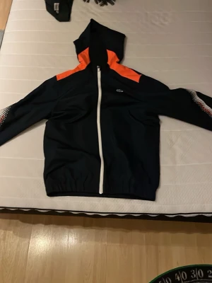 Lacoste vindjacka  - Snygg svart vindjacka från Lacoste med huva, vita dragkedjor och orange detaljer på axlarna. Ärmarna har ett rutigt mönster i vitt och rött. Perfekt för en sportig och trendig look.