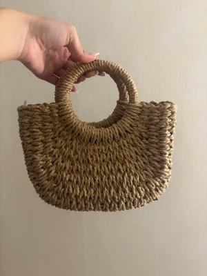 Flätad handväska i strå med runda handtag - Snygg handväska i naturfärgat stråmaterial med rund form och matchande runda handtag. Perfekt accessoar för en trendig och avslappnad look. Väskan har en bohemisk känsla och passar till många olika outfits. Oanvänd 