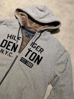Grå hoodie från Tommy Hilfiger - Snygg grå hoodie från Tommy Hilfiger med dragkedja och broderad text på bröstet. Insidan är fodrad med mjuk, beige fleece som gör den extra skön. Klassisk huva med dragsko och kängurufickor framtill. Perfekt för en chill och avslappnad stil.