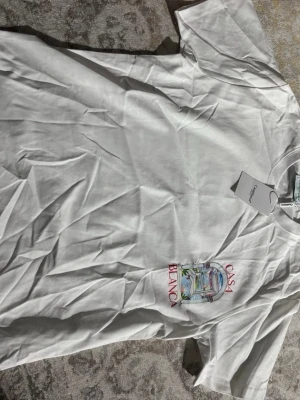Vit t-shirt från Casablanca - Vit t-shirt från Casablanca med färgglad grafisk print på både bröstet och ryggen. Trycket visar ett konstnärligt fönster med utsikt och texten 'CASABLANCA' i rött. Klassisk passform och rund hals. Perfekt för dig som gillar stilrena men unika plagg.