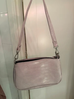 Rosa/lila handväska i crocopräglat - Trendig ljuslila handväska i crocopräglat konstläder med avtagbar axelrem och silverfärgade detaljer. Väskan har en stilren, rektangulär form och stängs med dragkedja upptill. Perfekt accessoar för att lyfta din outfit.