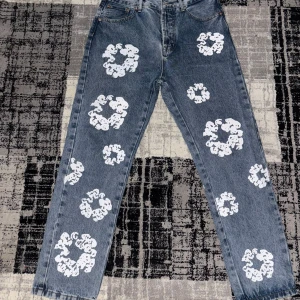 Denim Tears jeans  - Denim Tears jeans – Regler Blå jeans från Denim Tears med deras ikoniska vita cotton wreath-tryck över hela byxorna. Klassisk jeansmodell med rak passform och fem fickor. Ett stilrent streetwear-plagg som sticker ut.