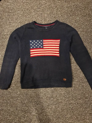 Mörkblå stickad tröja USA-flagga - Mörkblå stickad tröja från GANT med en stor amerikansk flagga framtill. Tröjan har rund halsringning, långa ärmar och ribbstickade muddar. Perfekt för dig som gillar klassisk och sportig stil.