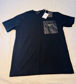 Svart t-shirt från Prada med ficka - Snygg svart t-shirt från Prada med en unik bröstficka i nylon och den klassiska metalltriangeln. T-shirten har rund hals och korta ärmar, perfekt för en clean och exklusiv look. Materialet är mjuk bomull med en detaljficka i nylon. Säljs då den aldrig används. 