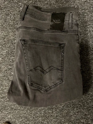 Grå jeans från Replay - Säljer ett par snygga grå jeans från Replay med klassisk femficksmodell och diskret broderad detalj på bakfickan. Jeansen har raka ben och svarta sömmar som ger en clean look. Perfekta till en avslappnad streetstil.