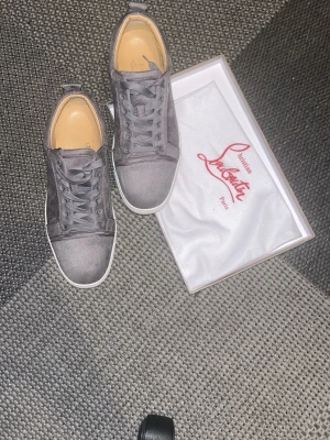 Gråa mockasneakers från Louboutin - Säljer ett par stilrena gråa sneakers från Christian Louboutin i mocka med vita sulor och klassisk röd detalj på undersidan, clean design som passar till det mesta. Storlek 42 men kan även passa 43