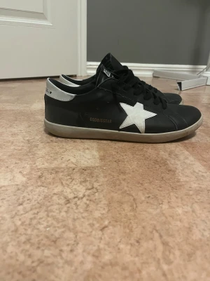 Golden Goose svart/vit sneakers - Svarta sneakers från Golden Goose med vit stjärndetalj på sidan och vit häl. Modellen heter Super-Star och har svarta skosnören samt en låg silhuett. Skorna har en klassisk design med gummisula och är tillverkade i läder.
