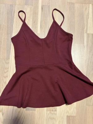 Vinröd peplum-linnetopp med smala axelband - Snygg vinröd peplum-linnetopp med smala axelband och v-ringning. Toppen har en figurnära passform upptill och ett utsvängt peplum nedtill. Perfekt att matcha med jeans eller kjol för en trendig look.