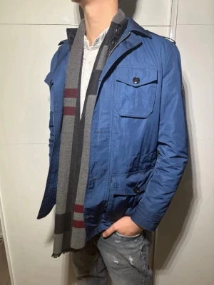 Blå vindjacka från j.lindberg - Snygg blå vindjacka från j.lindberg med klassisk krage, bröstfickor med knappar och axelklaffar. Jackan har en modern passform och diskret logotyp på ärmen. Perfekt för dig som vill ha en stilren och funktionell jacka till vardags.