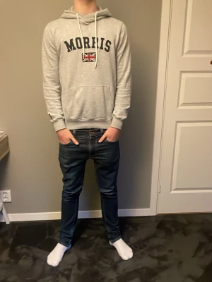 Grå hoodie från Morris med tryck - Snygg grå hoodie från Morris med svart text och brittisk flagga på bröstet. Klassisk känguruficka framtill och justerbar huva med vita snören. Perfekt för en avslappnad och stilren look. Passar folk mellan 170-185