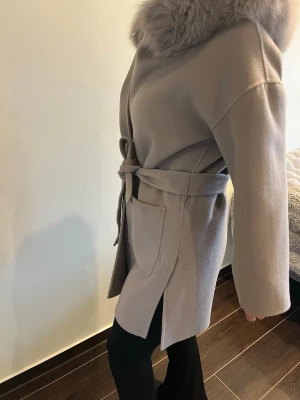 Beige kappa med äkta päls och knytskärp - Stilren beige kappa med bred äkta päls runt kragen och knytskärp i midjan. Kappan har stora fickor framtill och rak passform. Perfekt för dig som vill ha en trendig och mysig look under kalla dagar.