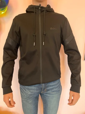 Svart hoodie från BOSS med dragkedja - Snygg svart hoodie från BOSS med dragkedja framtill och justerbar huva med snören. Säljer då jag inte har användning för dne längre den är i bra skick och tunn så bra till vår och sommar 