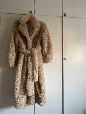 Beige lång teddyjacka med bälte - Supermysig beige teddyjacka i lång modell med bred krage och knytskärp i midjan. Jackan har fluffig struktur och vida ärmar, perfekt för kalla dagar när du vill vara både varm och snygg. -L: 120 cm -Ny men inte med prislapp på. Aldrig använd 