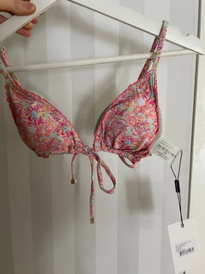 Blommig bikiniöverdel från By Malina - Söt bikiniöverdel från By Malina med färgglatt blommigt mönster i rosa, blått och grönt. Triangelformad modell med knyt framtill och justerbara axelband. Perfekt för sommarens stranddagar. Oanvänd!!!