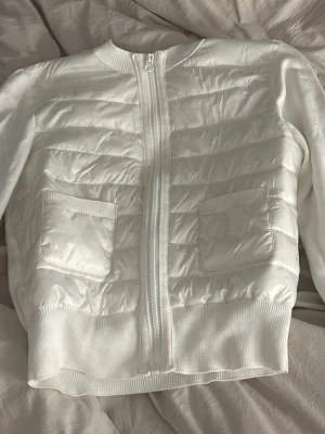 Vit quiltad cardiganjacka med fickor - Snygg vit cardiganjacka med quiltad framsida och två praktiska fickor. Jackan har dragkedja och ribbade muddar vid ärmslut och nederkant. Materialet är en mix av stickat tyg och quiltad polyester, vilket ger en modern look.