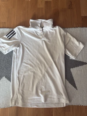 Vit pikétröja från Adidas (golf) - Snygg vit pikétröja från Adidas med klassisk krage och knäppning framtill. Tröjan har korta ärmar och tre svarta ränder på vänster ax sopm ger en sportig look. sopm ger en sportig look ram. Tillverkad i mjukt och lätt material, begärd för träning eller casual. (Golf) material