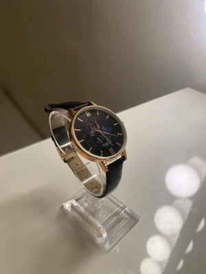 Svart klocka med rosdetalj - Stilren klocka med svart läderband och urtavla i svart med en ros i lila och blått. Boetten är i guldton och visarna matchar med guldfärg. Perfekt accessoar för dig som vill ha något unikt på handleden.