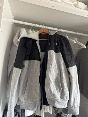 Ralph Lauren jacka  - Grå soprtig cardiganjacka från Polo Ralph Lauren med svarta quiltade partier över axlar och brish, dragkedja framtill och huva. Jackan har två fickor och den klassiska loggan broderad på bröstet. Perfekt för en avslappnad streetwear-look. Passar perfekt till vår, sommar och höst. M men mer åt S hållet 