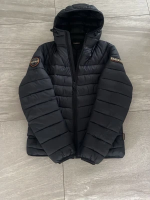 Svart dunjacka från Napapijri - Snygg svart dunjacka från Napapijri med huva och dragkedja framtill. Inprincip helt ny, andvänd två gånger. Passar perfekt för mig som 175 lång. 