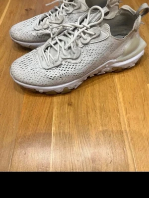 Nike React vision 55  - Snygga Nike React vision 55 sneakers i beige mesh med vita och ljusbruna detaljer. Skorna har en sportig design med ventilerande ovandel, snörning och en unik, vågig sula som ger grym komfort. Perfekta för dig som gillar stilrena och lätta sneakers.