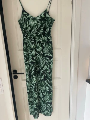 Grön klänning  med tropiskt mönster - Snygg klänning med smala axelband och tropiskt bladmotiv i olika gröna nyanser. Lång modell med markerad midja och v-ringad hals. Slits på båda sidorna. Perfekt för dig som vill sticka ut med en fräsch och somrig look. Materialet känns lätt och luftigt.