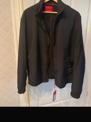 Svart jacka från HUGO med dragkedja - Stilren svart jacka från HUGO med klassisk krage och hel dragkedja framtill. Jackan har släta fickor på sidorna och diskret röd HUGO-detalj vid dragkedjan. Materialet är mjukt och slätt, ger en clean look. Helt ny. Passar M-L, storlek 54 Hugo. Köpte förra hösten för 2990:- men tyvärr för liten nu.