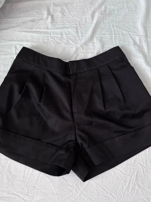 Svarta dressade shorts med hög midja - Snygga svarta shorts med hög midja och uppvikta benslut med 2 sido fickor. De har pressveck framtill, dold dragkedja och hyska/hake-stängning samt stretchande baktill. Materialet är semi-glansigt. Passar storlek s/m, de står xxl innuti men de är köpa i Asien så därför är de extremt mycket mindre i storleken. Skön och lite mer strukturerad. Aldrig använda de va för små