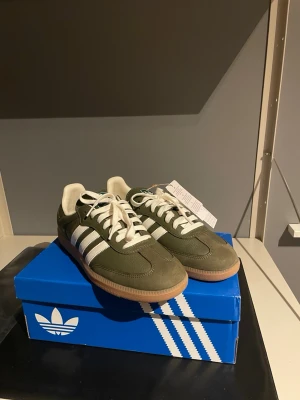 Adidas Samba OG gröna sneakers - Adidas Samba OG sneakers i olivgrönt läder och mocka med klassiska vita ränder och vit häl. Skorna har gummisula och vita skosnören. Perfekt för dig som gillar retrostil och streetwear. Modellen har låg profil och guldfärgad Samba-text på sidan. Aldrig använda och prislappen är kvar. Hör av dig vid mer frågor!