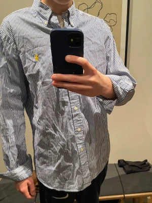 Blåvit randig skjorta från Ralph Lauren - Säljer en stilren blåvit randig skjorta från Ralph Lauren i klassisk passform. Skjortan har knappar framtill, button-down krage och den ikoniska gula Polo-loggan broderad på bröstet. Perfekt för dig som gillar stilrena och tidlösa plagg. Skjortan är i väldigt bra skick. Ny pris 1599 kr. Hör av dig vid funderingar!