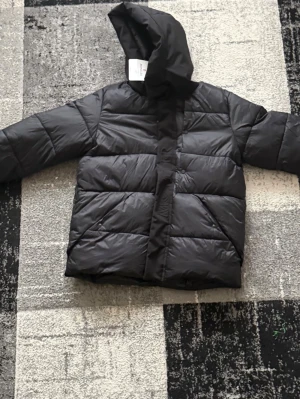 Svart dunjacka med huva från Moncler - Säljer en svart dunjacka från Moncler med stor vadderad huva och breda quiltade paneler. Jackan har dold dragkedja och tryckknappar framtill samt diskret Moncler-logga på knäppningen. Perfekt för kalla vinterdagar.