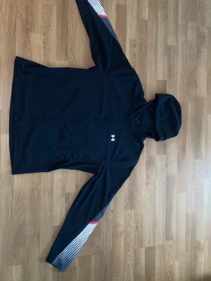Svart hoodie från Under Armour - Snygg svart hoodie från Under Armour med deras logga på bröstet. Hoodien har långa ärmar med vita och röda detaljer på ena ärmen. Tillverkad i ett lätt och ventilerande material som passar perfekt för träning eller chill.