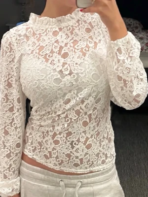 Vit spetsblus med hög krage - Säljer en superfin vit spetsblus med lång ärm och hög krage. Blusen har ett vackert blommigt spetsmönster, transparent material och knappdetalj i nacken. Perfekt för dig som gillar romantisk och trendig stil.