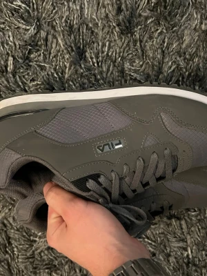 Grå sneakers från Fila - Snygga grå sneakers från Fila med klassisk siluett och vita sulor. Skorna har meshdetaljer på sidorna för extra andningsförmåga och är tillverkade i en mix av syntetmaterial och textil. Fila-logga på sidan och plös, samt matchande grå skosnören.