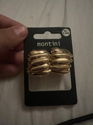 Guldiga chunky örhängen Montini - Säljer ett par chunky örhängen i guldig metall från Montini. De har en vågig, modern design som verkligen sticker ut och ger en cool touch till din look. Perfekta för dig som gillar statement-smycken och vill ha något som syns.