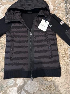 Svart moncler cardigan - Snygg svart pufferjacka från Moncler med huva och ribbade muddar. Jackan har quiltad front, två dragkedjeförsedda fickor och Moncler-logga på ärmen. Ärmarna är stickade för extra komfort och stil. Perfekt för året om. Om du har några funderingar eller frågor ställ dem gärna 